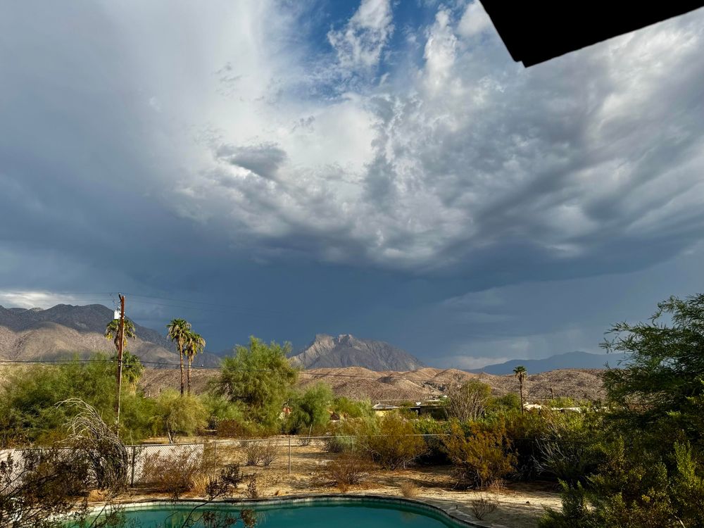 iPhone wide shot taken from our back porch featuring gorgeous dark rain clouds above the grey and brown and purple mountains with desert vegetation all around and a swimming pool in the foreground