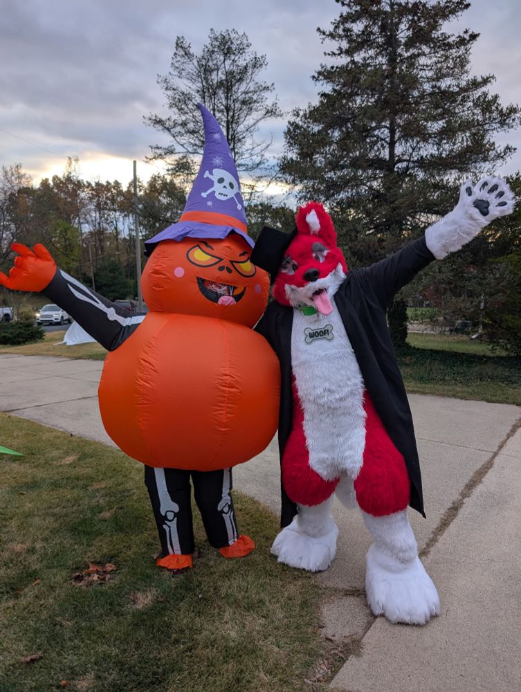 A pumpkin inflatable next to a red and white doggo fursuiter posing for the camera.