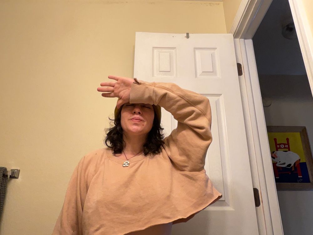 Left arm over head symbolizing healing and unleashing of creativity during times of struggle. Location: personal bathroom with yellow painted wall and white door. Wearing necklace from lover, crop top sweater and skull cap