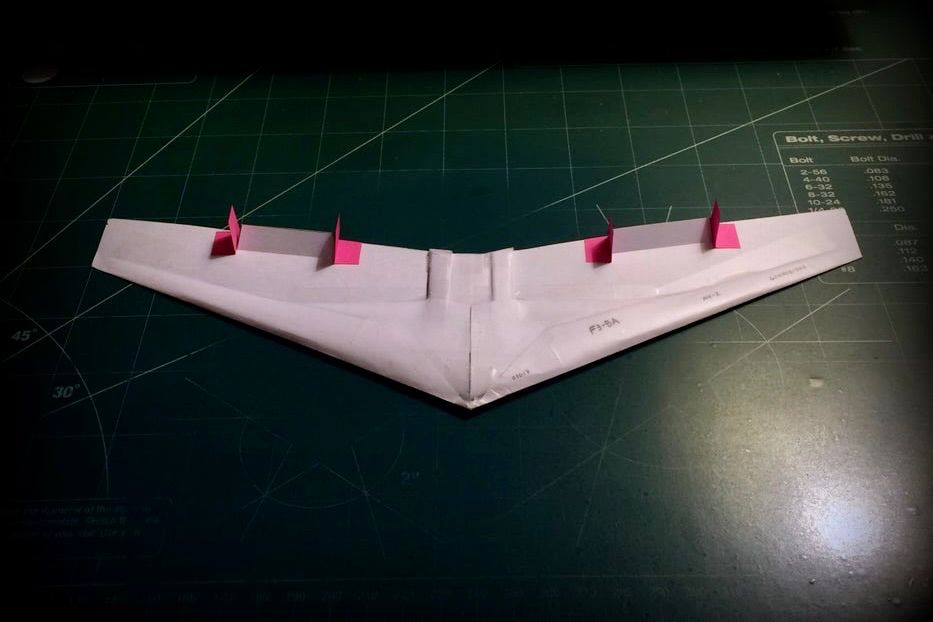 A small paper model of a flying wing vaguely resembling Jack Northrop's YB-49, at least in terms of its generalised layout. The model is viewed directly in front of and slightly above, 'flying' directly towards the camera position. Along the trailing edge of the wing, there are four small pink pieces of paper formed into vertical stabilisers, also similar to those found on the YB-49. The rest of the aircraft is primarily white. The aircraft is sitting on a green, self-healing cutting mat. Original caption from source: « How to Make the Omniwing Swift Paper Airplane » (📸 OrigamiAirEnforce via Instructables under CC BY-NC-SA 4.0)