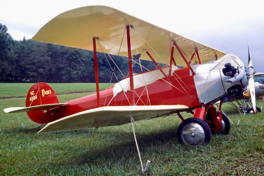 A taildragger biplane is parked and secured in a smooth, green, grassy field. The aircraft is oriented pointing to the right such that the right side of the aircraft is visible almost in profile. The wings and horizontal stabiliser are a cream colour, whereas the fuselage and vertical stabiliser are bright red. The registration, « NC499H », is painted on the rudder. The Parks logo is painted on the vertical stabiliser. There is a radial engine with the cylinders exposed to the slipstream. The fuselage just aft of the engine is sheathed in aluminum. The cockpit is covered with a tarp indicating flight is not imminent. A line of leafy trees is visible in the distance. The sky appears to be grey overcast.  Original caption from source: « Detroit-Parks P-2A ¶ One of eleven built in 1929-30, on display at the EAA Fly-In. This is the airplane flown by Richard Bach in the book "Nothing by Chance", his account of barnstorming in the US during the late 1960s. ¶ Tullahoma - Regional / William Northern Field (THA) ¶ Tennessee, USA - September 29, 1979 » (©1979, 2025 David Lednicer, use with his kind permission.)