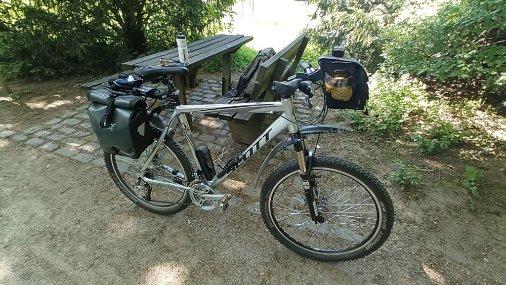 Kein Problem! Möchtest du eine Bildbeschreibung, die sachlich ist, oder lieber eine kreative oder stimmungsvolle Version? Hier ist eine sachliche Variante als Startpunkt:

Bildbeschreibung:

Das Bild zeigt ein voll ausgestattetes Tourenrad der Marke Scott, das an einem schattigen Rastplatz im Wald steht. Am Fahrrad sind mehrere Gepäcktaschen angebracht – eine große Tasche am Gepäckträger hinten sowie eine Lenkertasche vorne, die geöffnet ist und Proviant enthält. Auf dem Tisch im Hintergrund stehen eine Trinkflasche und eine Sonnenbrille, daneben liegt ein Rucksack auf der Bank. Der Weg besteht aus festem Sandboden, daneben ist ein Abschnitt mit Pflastersteinen. Rundherum wachsen dichte grüne Sträucher und Bäume, die eine angenehme, naturverbundene Atmosphäre schaffen.

