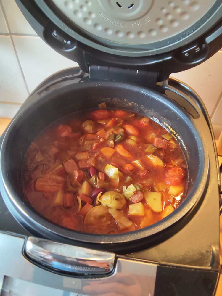 **Deutsch:**
Das Bild zeigt einen Topf mit einem Eintopfgericht, das verschiedene Zutaten wie Bohnen, Kartoffeln, Tomaten, Paprika und Würstchen in einer roten, würzigen Soße enthält. Der Topf befindet sich in einem Multikocher oder einer Küchenmaschine.

**English:**
The image shows a pot with a stew containing various ingredients such as beans, potatoes, tomatoes, peppers, and sausages in a red, spicy sauce. The pot is inside a multi-cooker or kitchen appliance.