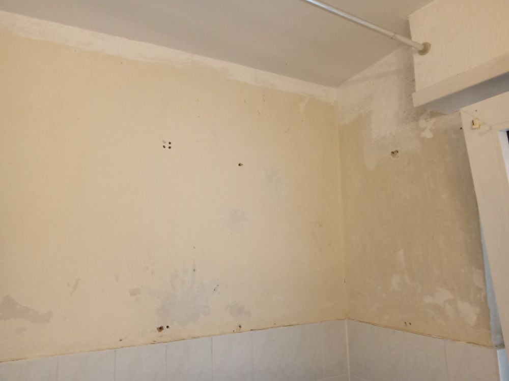 Das Bild zeigt eine Innenwand eines Raumes, vermutlich eines Badezimmers oder einer Küche. Die Wand weist verschiedene Schäden auf, darunter Verfärbungen, Flecken, Unebenheiten und kleine Löcher. Die obere Hälfte der Wand ist beige oder hellgelb gestrichen, während die untere Hälfte mit weißen Fliesen verkleidet ist. In der rechten oberen Ecke befindet sich eine hervorstehende Konstruktion mit einer weißen Stange, möglicherweise eine Duschvorhangstange. Die Wand scheint Spuren von Feuchtigkeit oder unvollständig entfernter alter Farbe zu zeigen.