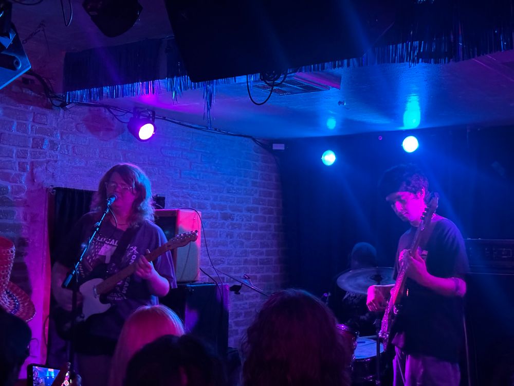 The three piece rock band “Proun” on stage at the Chess Club in Austin, TX, on May 9th 2025. The lighting is a mix of blue and pink.