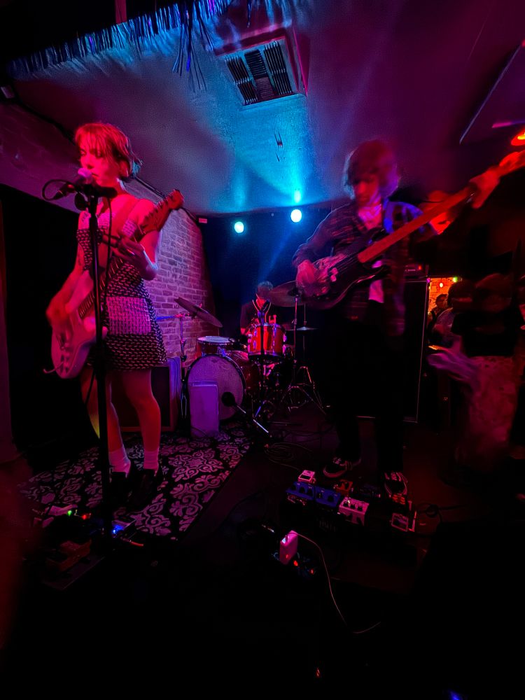 Three piece rock band flooding, performing at Chess Club in Austin TX on May 9, 2025. A wide angle photo, from the front row. The lead singer is mostly in focus, the bass player are blurred from motion.