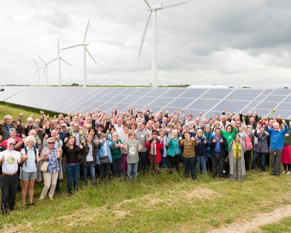 A large gathering of people in front of the Westmill solar farm, with wind turbines in the background.