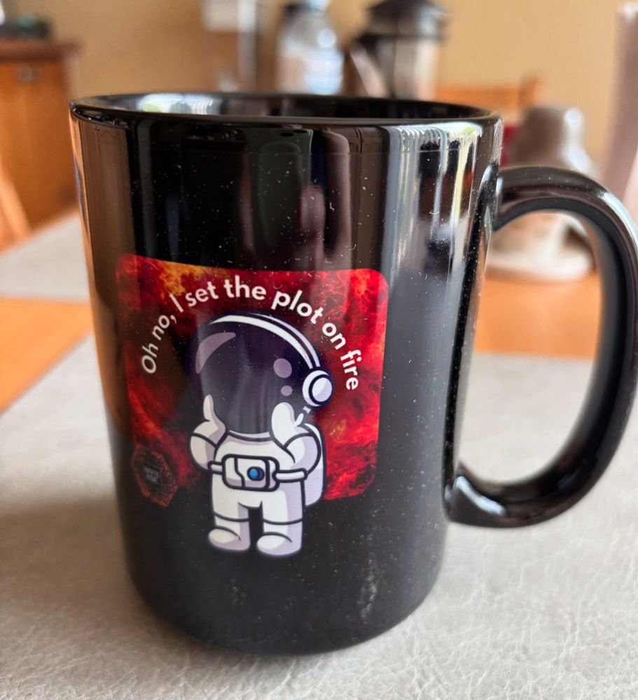 A black mug, with a little astronaut grasping their face while surrounded by fire. Text says: Oh no, I set the plot on fire.
