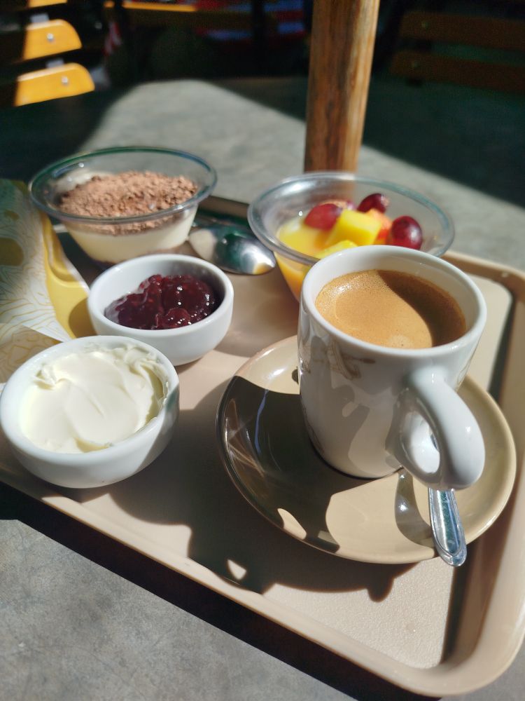 Foto de uma café da manhã do Sesc, tem uma xícara com café e um pires, dois potinhos na esquerda com manteiga e geleia, um doce no canto esquerdo e uma salada de fruta no canto direito.