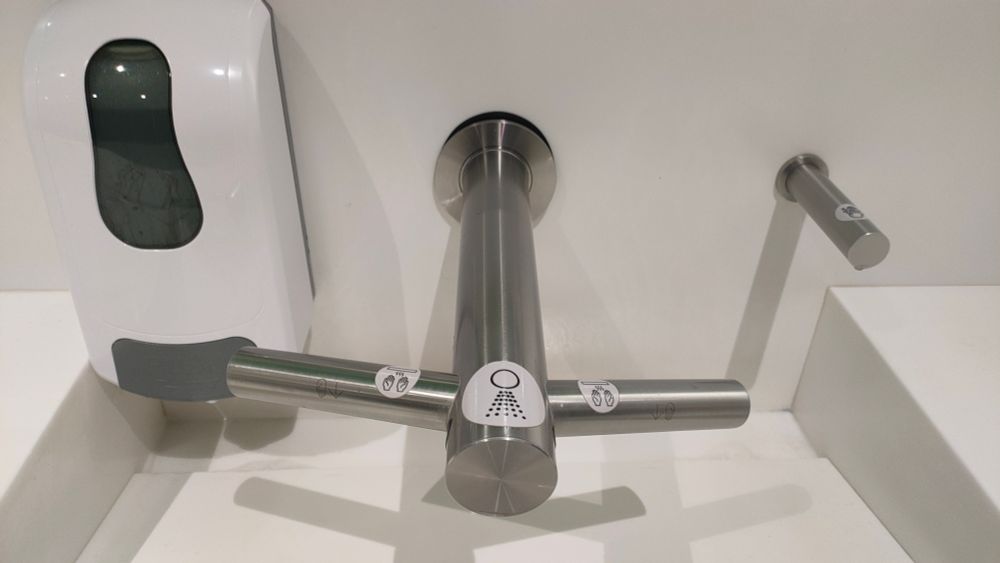 A public bathroom sink in which the dryer has been incorporated into the tap. To the right is a matching soap dispenser projecting straight from the wall, there is no obvious way to refill it. On the left is the standard soap dispenser that has been installed afterwards because no one can be bothered filling up the one on the right.
