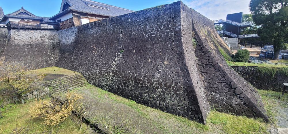 A photo of the retaining wall around Kumamoto Castle, made of huge black stones. The wall swoops gently downwards instead of going in a straight line. 