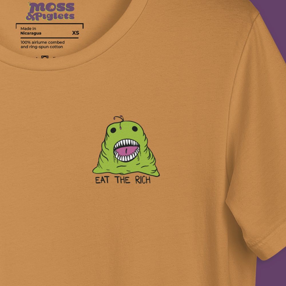 A close up a tshirt design. The design is a green slime monster with an open mouth and tons of teeth. Drool is coming out of the mouth and underneath the design it reads “eat the rich”