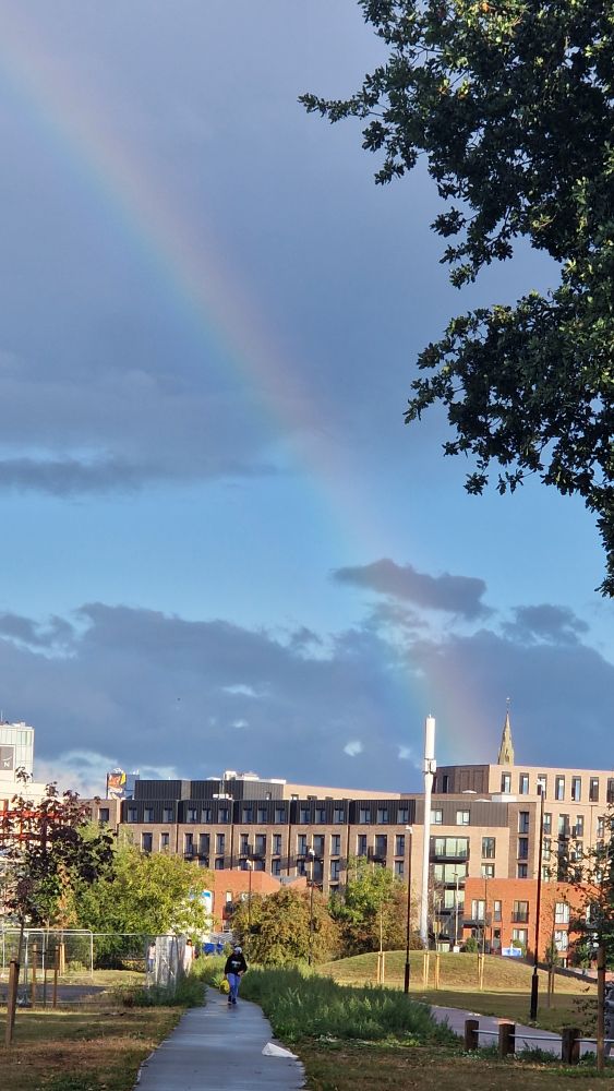 Again and portrait image. A path cross grass. Then buildings and behind them a spire peeks out. A Rainbow goes from it diagonally to the left