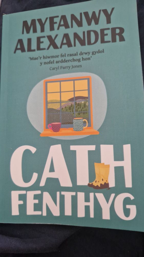 Photo of Cath Fenthyg by Myfanwy Alexander.  It is bluey green with the author's name in black capitals at the top and the title in white capitals at the bottom.  In the musky is a drawing of a window looking out on a hilly landscape and two mugs on the window sill.  In the TH of Cath there is a drawing on yellow wellies with mud on the bottom.  Under the author's name is a recommendation from Caryl Parry Jones "Mae'r hiwmor fel rasal drwy gydol y nofel ardderchog hon" 