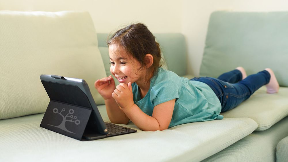 Young girl lying on a couch, smiling at a tablet with a keyboard case, suggesting she's engaged in watching or interacting with content.