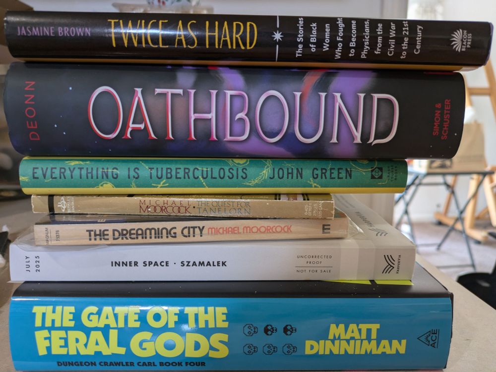 A stack of books, from top to bottom: Twice as Hard by Jasmine Brown, Oathbound by Tracy Deonn, Everything is Tuberculosis by John Green, The Quest for Tanelorn by Michael Moorcock, The Dreaming City by Michael Moorcock, Inner Space by Jakub Szamalek, and The Gate of the Feral Gods by Matt Dinninan.