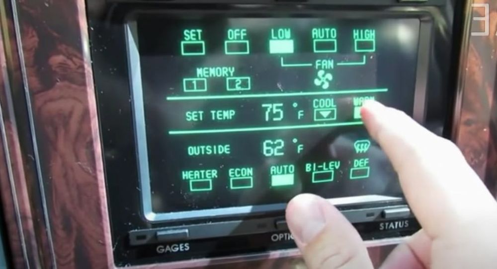 Monochrome touchscreen monitor with bright green icons for vehicle controls, with a fake burled-woodgrain border, in a 1986 Buick Riviera.