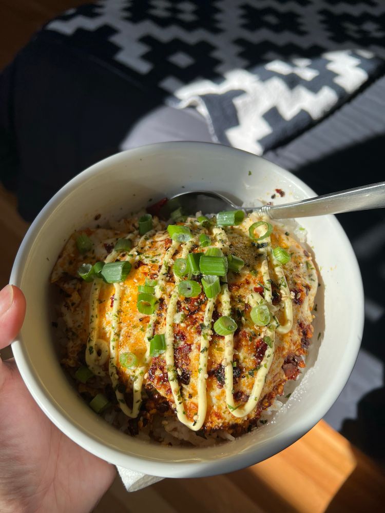 yuzu’s hand holding a white ceramic bowl filled with white jasmine rice and two chili crisp fried eggs, topped with kewpie mayo, furikake, and green onions; a silver spoon rests on the inside of the bowl. hardwood floors, black blankets, and a black and white throw designed with a pattern of symmetrical shapes can be seen in the background.