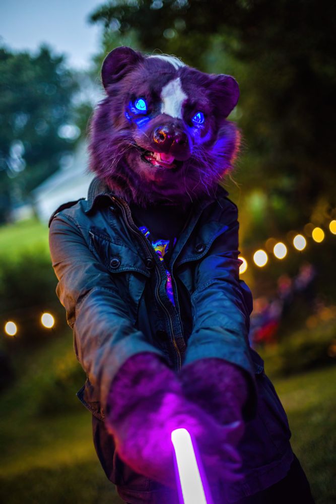 A person wearing a skunk head costume and weathered jacket hold a purple light in their paws out in front of them. Their eyes glow blue. They are standing in a backyard with a string of lights hung behind them.