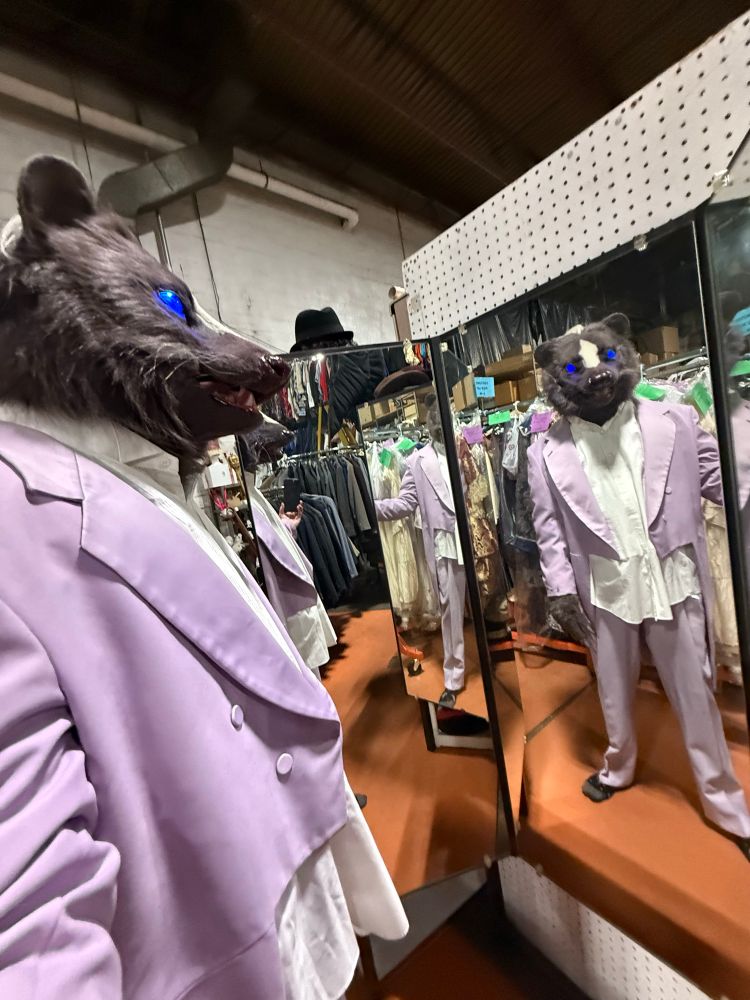 A person wearing a skunk head costume stands in front of a multipart mirror. They are trying on a light purple tuxedo.