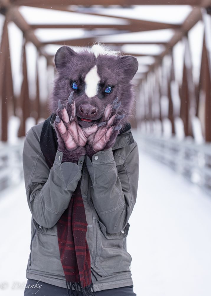 A person in a skunk head costume holds their paws to their face. They are standing in front of a metal bridge in winter with snow covering the ground. They are wearing a grey jacket and a red scarf.