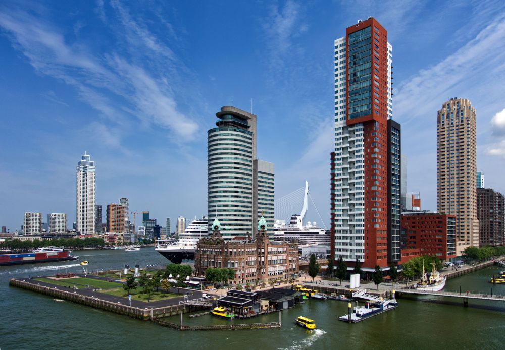 Foto van de Kop van Zuid en de skyline van Rotterdam, met een containerschip dat door de Maas vaart en een cruiseschip van de HAL die Rotterdam heet