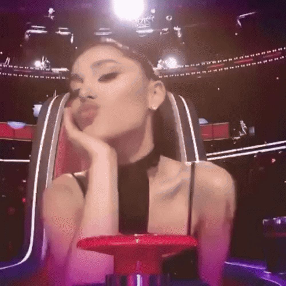 ariana grande is sitting in a chair with her hand on her chin and blowing a kiss .