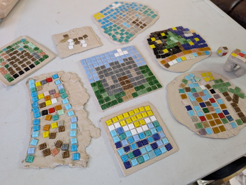 Aerial photo of mosaic creations on a table. The designs include 2 castles, a starry night with a tree, and a beach. The mosaics were created using colourful square glass tiles pushed into air dry clay. 