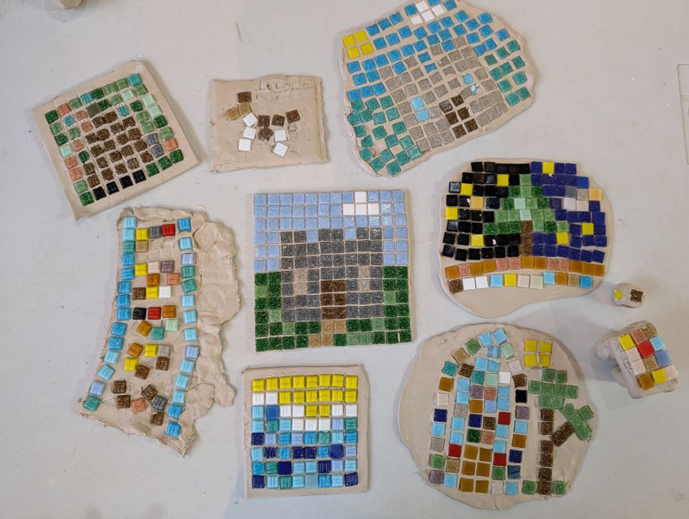 Aerial photo of mosaic creations on a table. The designs include 2 castles, a starry night with a tree and a beach. The mosaics were created using colourful square glass tiles pushed into air dry clay. 