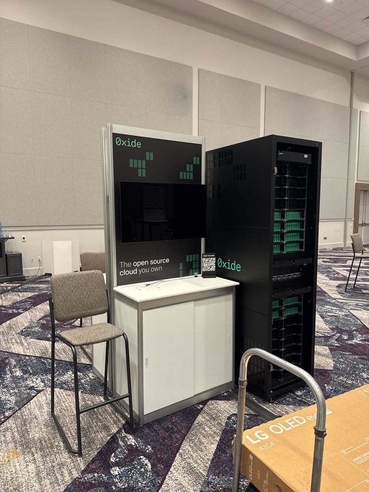 A photograph of an Oxide exhibit booth at SUSECON in a large conference hall. On the right stands a tall black server rack the Oxide cloud computer, featuring green accents in each bay. To the left is a freestanding sign with “Oxide” branding and the slogan “The open source cloud you own,”