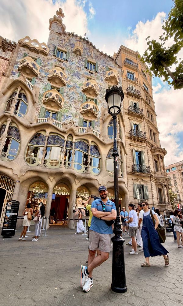 A picture of me in front of Casa Battló, an iconic building in Barcelona designed by Antoni Gaudí. I’m in the foreground wearing a blue polo and snazzy new sneakers, and the building facade behind me has smooth, flowing lines, warm concrete, and blue and green mosaic accents 