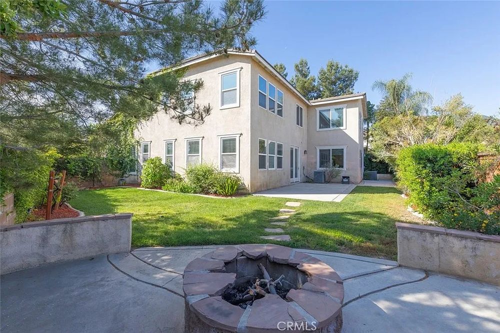 A picture of a relatively normal backyard with a weirdly shaped fire pit in the foreground. There are shadows cast on the lawn by trees behind the camera. The top lip of the pit is lined with stone stone wedges of a uniform width, but varying lengths. The difference of lengths is down to the fact they all appear to be broken, with the ends closest to the fire appearing shattered rather than cut. One wedge is completely missing a point, and others are failing to cover the concrete substructure. Nothing about this looks intentional.