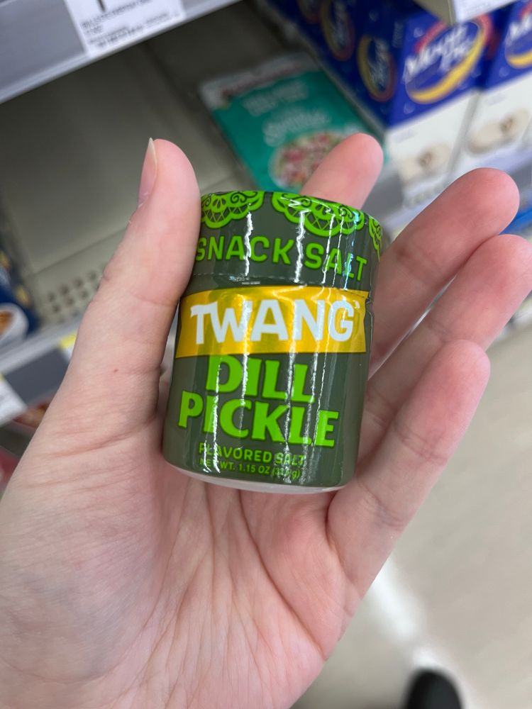 A pale hand holding a container of Twang brand snack salt in the flavor ‘dill pickle’