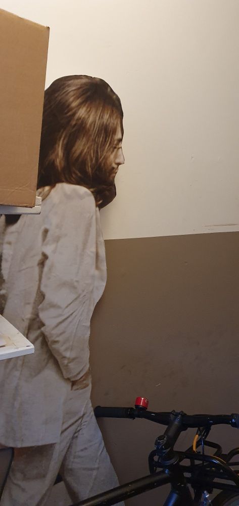 Life-size cardboard cut-out of John Lennon from the album cover of Abbey Road. Leaning on a wall in a hallway behind a bicycle and party covered by a shelf. 
