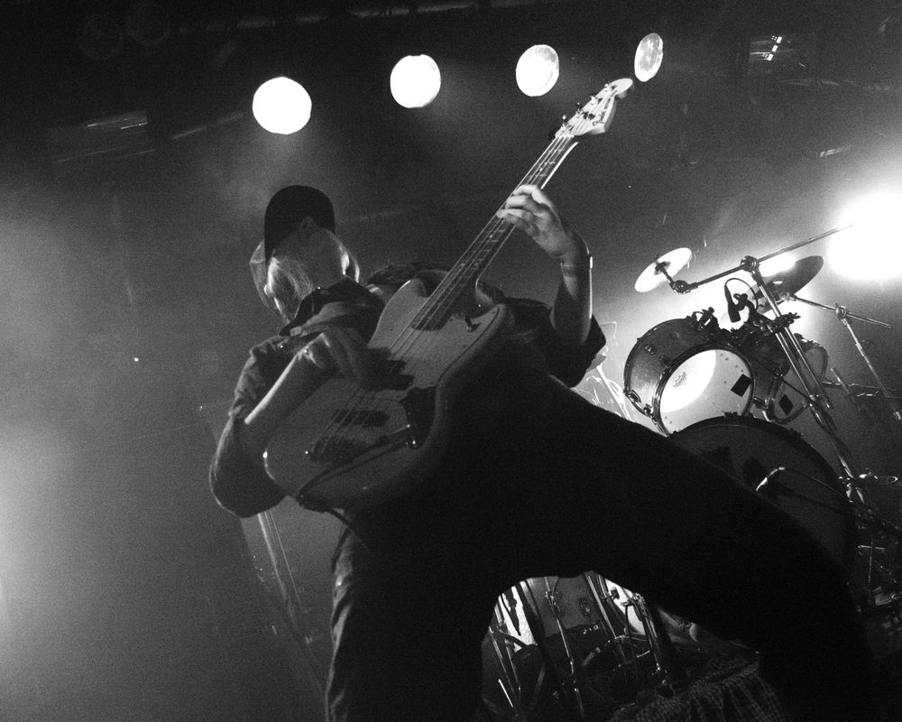 Black and white landscape photo of female bass player wearing a jumpsuit and a baseball cap, leaning back, legs wide apart, playing their bass guitar on a stage mid concert.