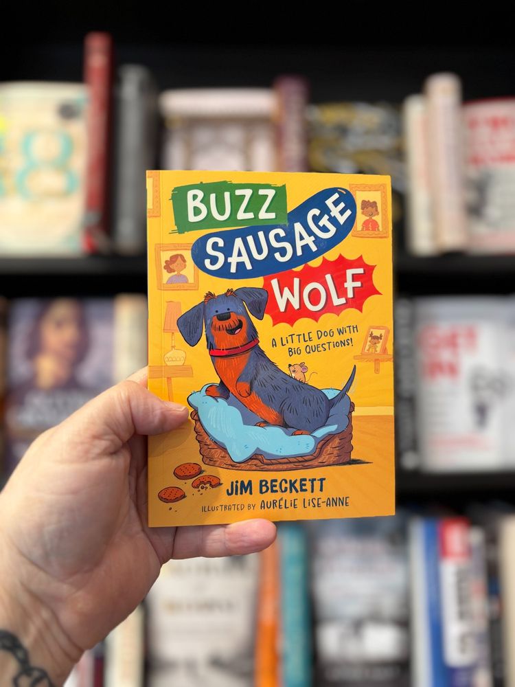 hand holding a book in front of a bookcase, on the book is a picture of a sausage dog in a dog bed