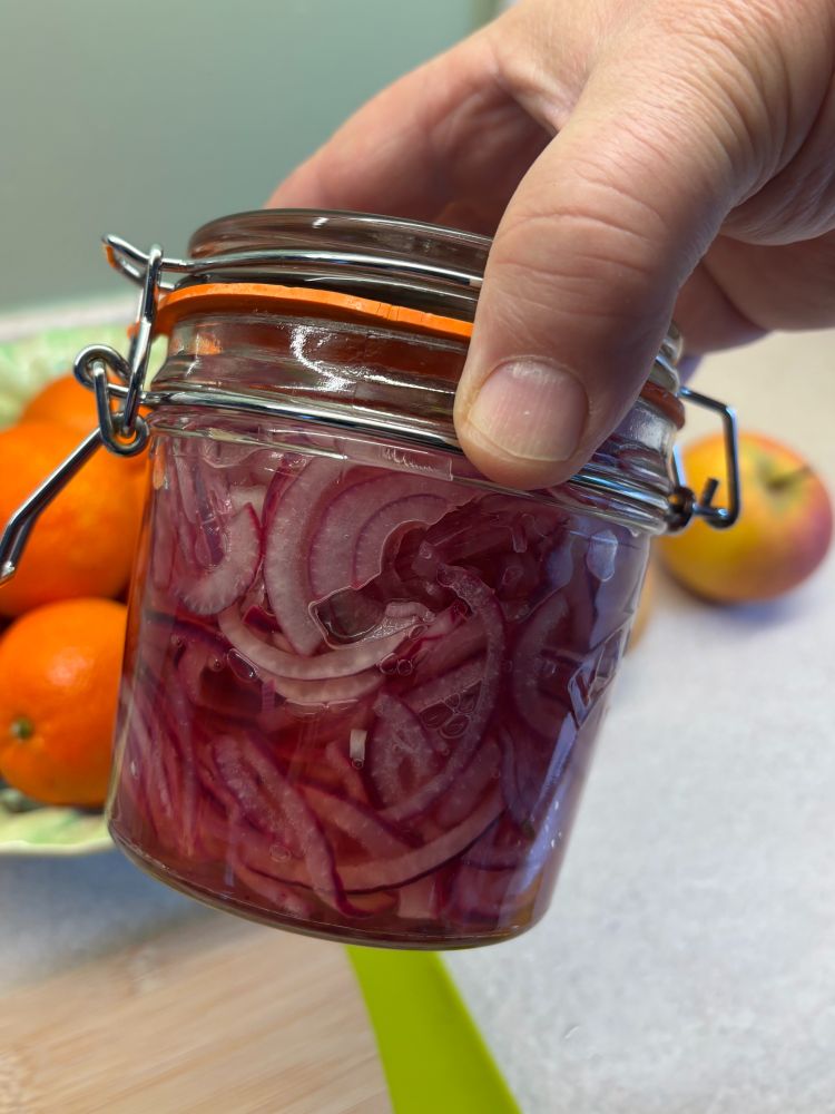 a hand holding a jar of pickled red onions behind the jar are some fruit and a chopping board