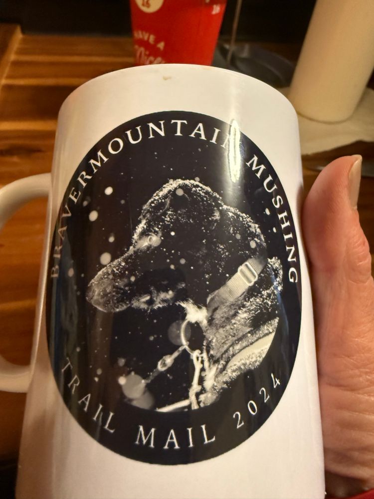 A coffee mug with the picture of a black sled dog, with a slight overbite, covered with a gentle snow. The cup reads BraverMountain Mushing Trail Mail 2024