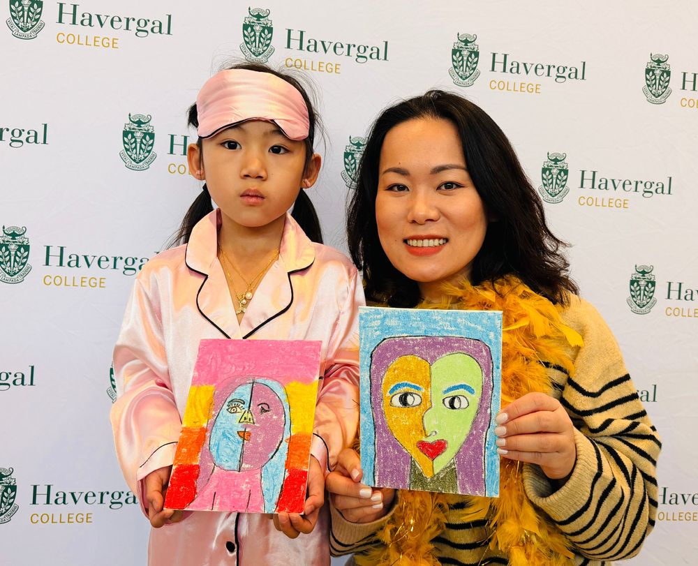 A Grade 1 student and her mom wearing pyjamas pose together while holding self-portraits in front of a white background with repeating Havergal College logos on it.