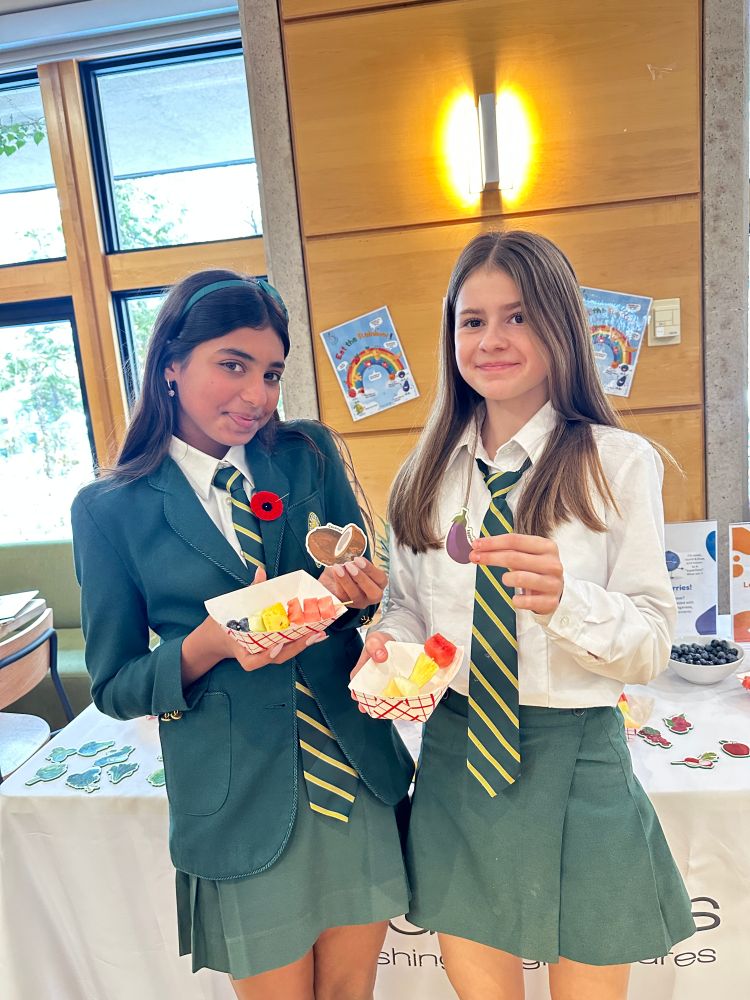 Two Upper School students pose together while holding fruit skewers and stickers depicting an eggplant and a coconut.