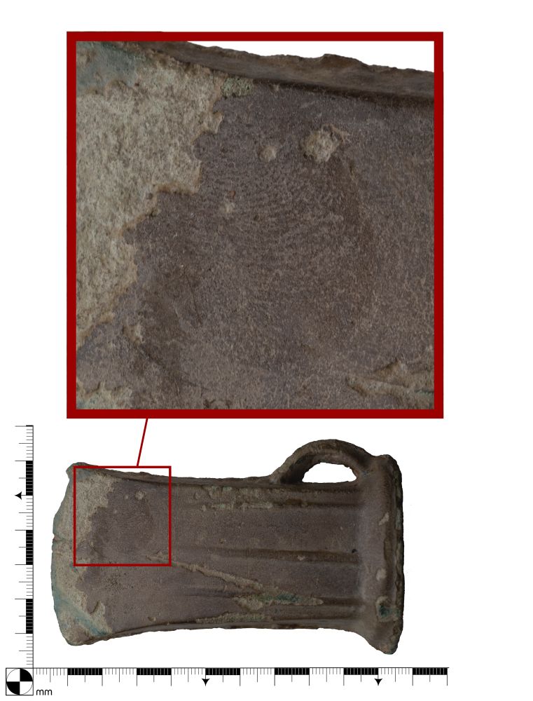 Photo of a Bronze Age socketed axe head with a zoomed in thumbnail of a finger print on one of the faces towards the blade edge. The axe is dark green with oxidation and the surface of the blade edge has been lost. 
