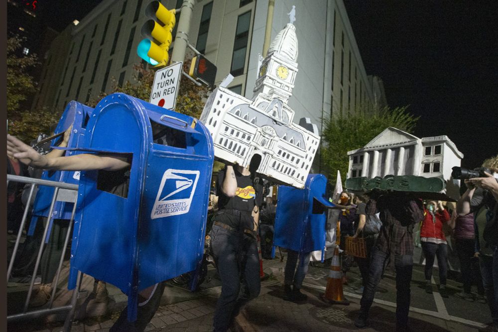 It's a little hard to describe so bear with me. It's nighttime, outside the Philadelphia convention center (during vote counting in November 2020). Some people are dancing, many more are in a crowd watching and taking pictures. Two dancers are dressed up like buildings, that is to say they're wearing normal clothes but covering their heads are enormous (like the size of a whole other person) recreations of the White House and Philadelphia City Hall. Three others are dressed up like blue USPS mailboxes you'd find on the street, including the most prominent dancer in the foreground who just has legs and arms sticking out and the mail slot at the top is open enough to see out of. The image source is this article about how one of the mailbox costumes is going in the Smithsonian: https://www.inquirer.com/arts/philadelphia-election-dancing-mailboxes-spiral-q-2020-20210708.html