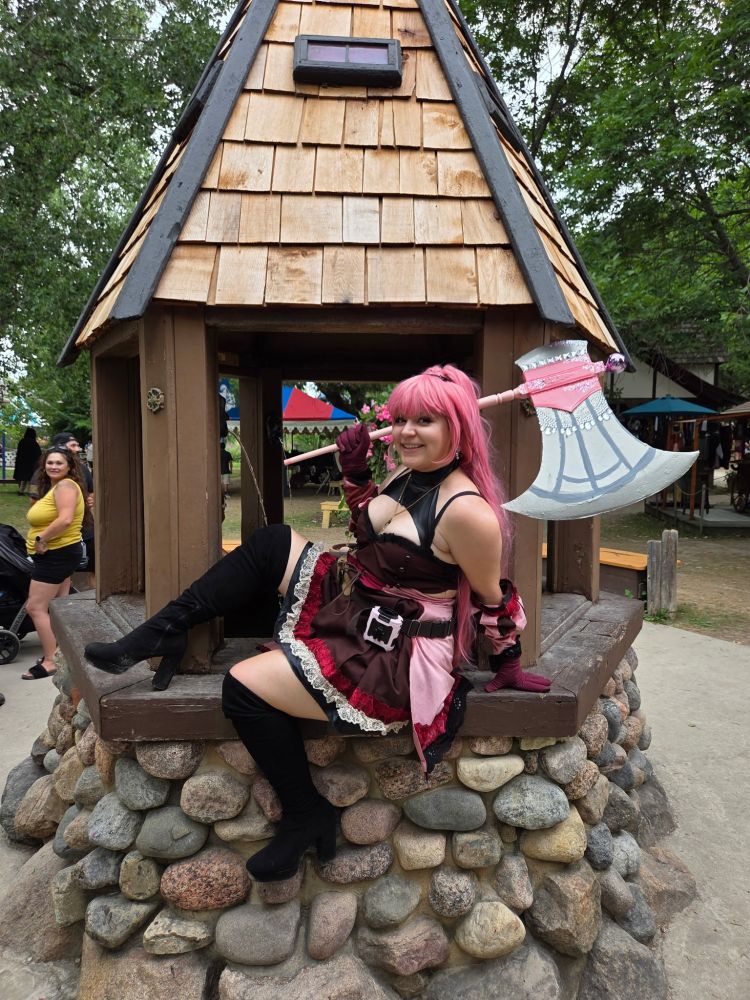 Photo of me cosplaying timeskip Hilda from Fire Emblem Three Houses. She has pink hair and a brown and pink dress and black boots. I'm sitting on the well near the entrance of the faire. I'm holding the axe I made myself that is NOT her relic weapon but instead, my own take on what Hilda would wield if she had the choice. A little girl made wide eyes at me as she saw me climb into the Well and said "that's not safe! But you look cool!"