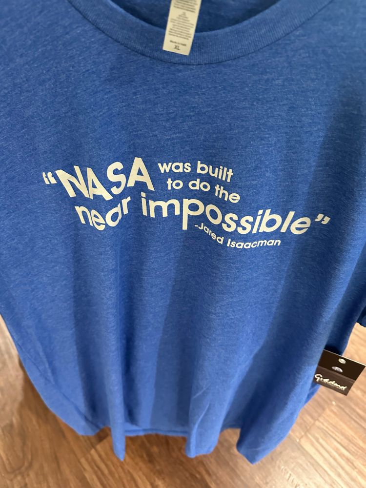 A blue shirt with the quote “NASA was built to do the near impossible” -Jared Isaacman
