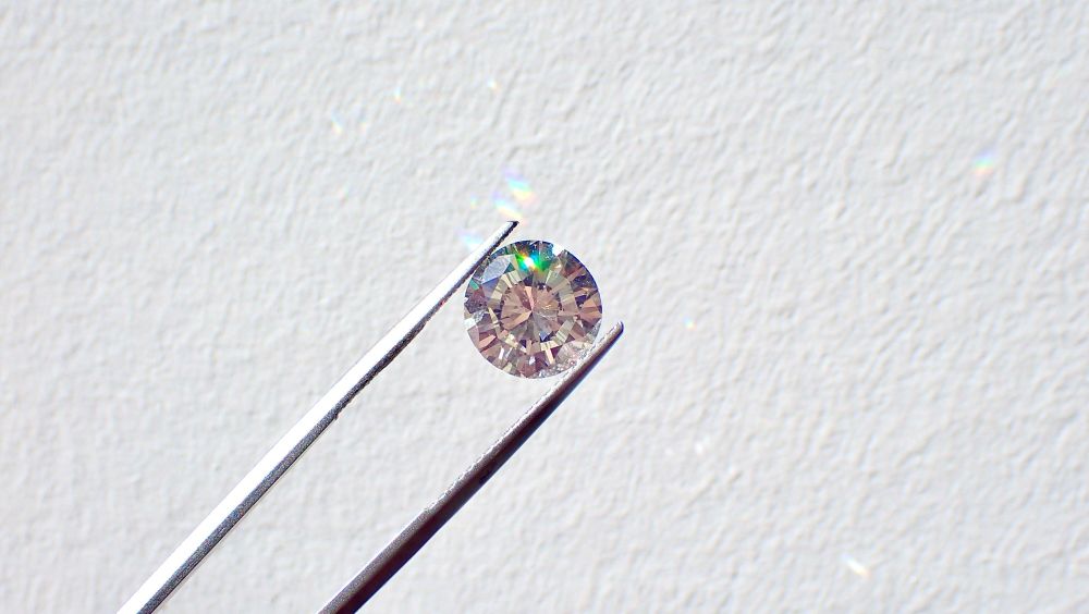 A diamond being held between a pair of silver tweezers so that it can be examined, over a plain white surface.