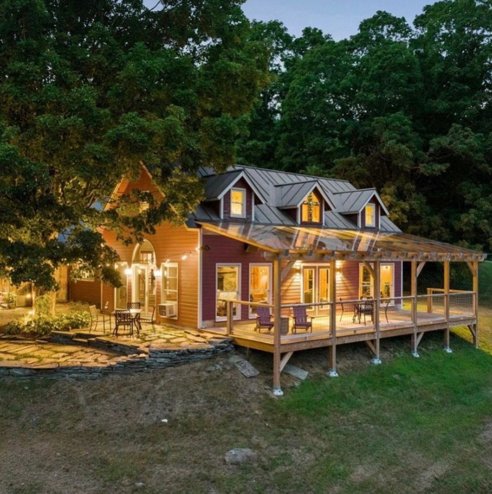 Red farmhouse lit up by glowing lights both inside and out. On the right is grass and a stone patio is on the left 