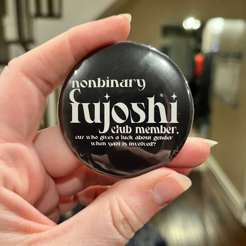 bendy holding a black button, and in white letters it says “non binary fujoshi club member.” under that it says “cuz who gives a fuck about gender when yaoi is involved.” 