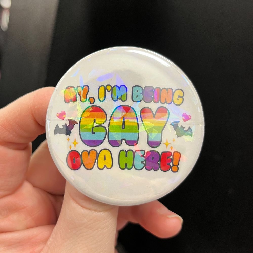 bendy holding a holographic button in rainbow colours that says “Ay, I’m being gay ova hear!” which is a quote that is from what we do in the shadows