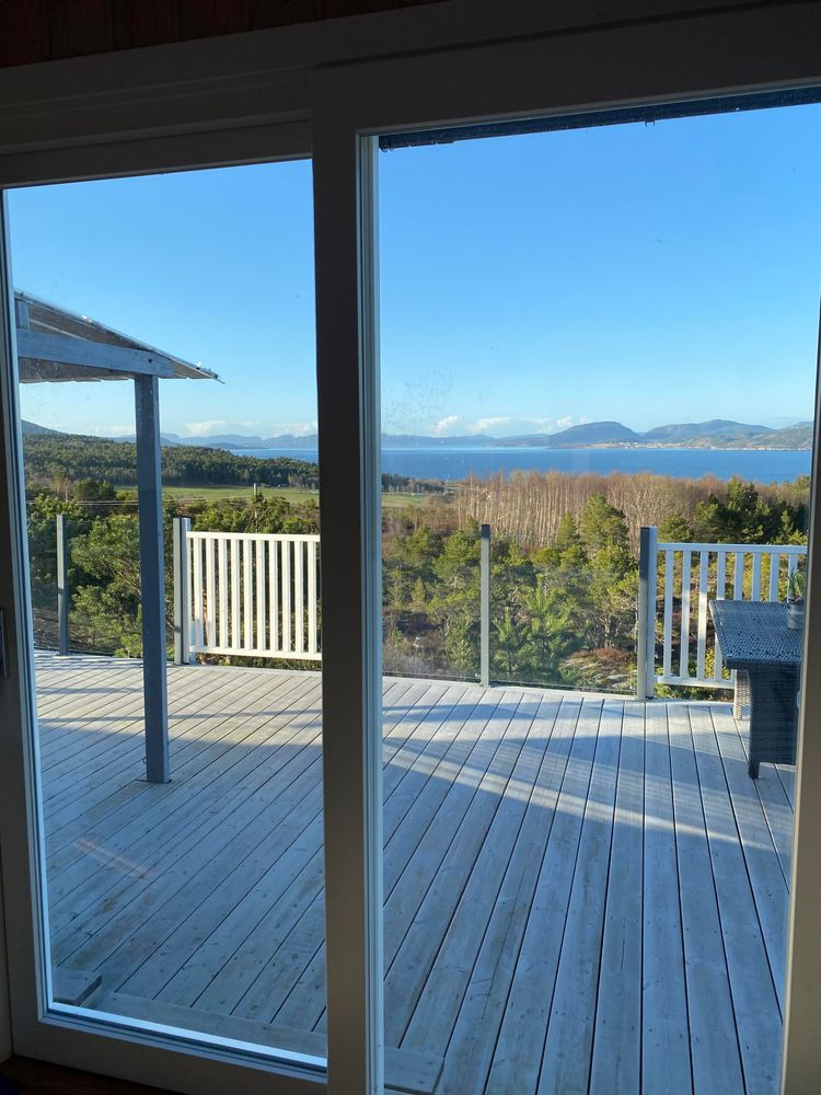 Utsikt mot terrassen og Trondheimfjorden gjennom skyvedørene på hytta. Strålende sol. Hvit terrasse med delvis glassrekkverk. Furuskog i forkant og blå-blå fjord og himmel ut gjennom Trondheimsleia.