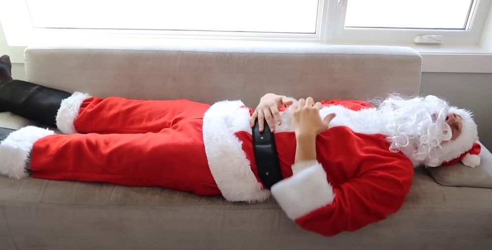 Santa Claus laid up on an IKEA couch grabbing his midsection like he ate a bad quiche